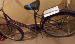 rower typu damka koloru fioletowego z tylnym  bagażnikiem i siodełkiem oklejonym niebieska taśmą